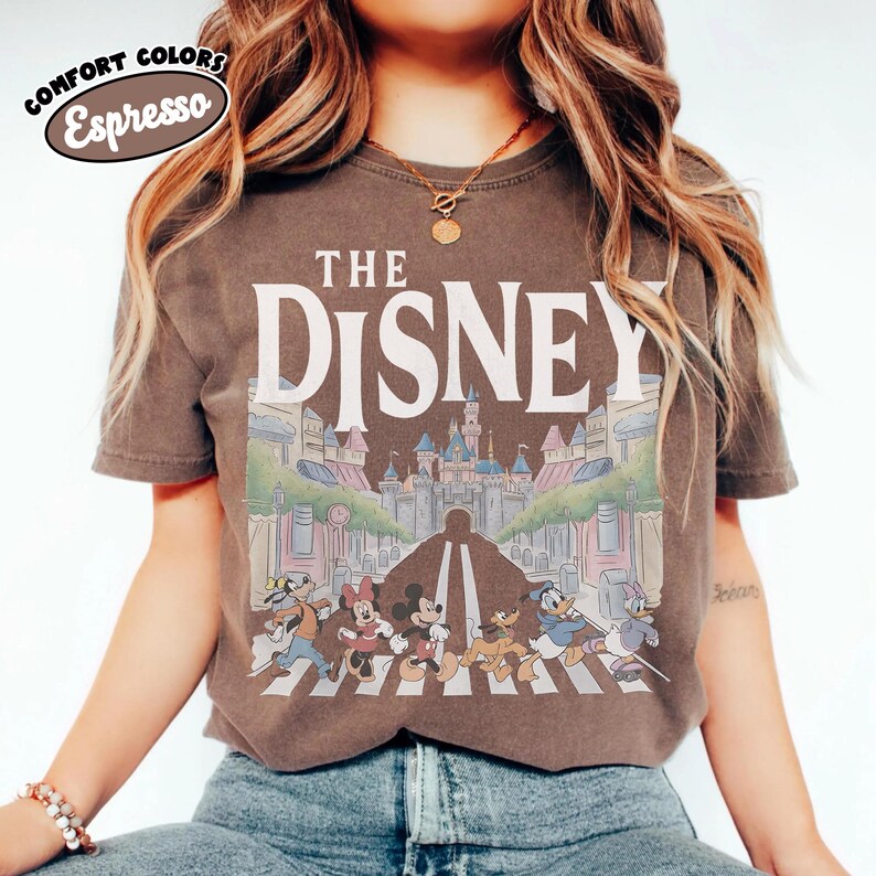 Może przedstawiać: Koszulka w kolorze espresso z napisem "THE DISNEY" nad kolorową ilustracją postaci Disneya przechodzących przez ulicę przed zamkiem. Koszulka ma wygląd vintage.