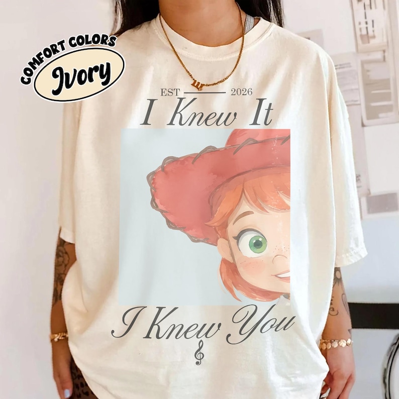 Op de afbeelding: Een ivoorkleurig t-shirt met een afbeelding van een stripfiguur met rood haar en een rode cowboyhoed. De tekst "EST 2026 I Knew It" staat boven de afbeelding en "I Knew You" met een muzieknoot eronder. Een logo toont "COMFORT COLORS IVORY".
