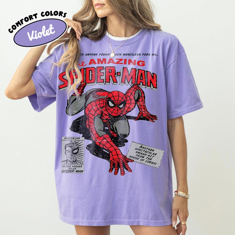 May include: A violet short-sleeve t-shirt featuring a distressed graphic of Spider-Man in a crouching pose. The text reads The Amazing Spider-Man, Featuring Peter Parker, and Another Spectacular Smash-Hit From The House Of Ideas.