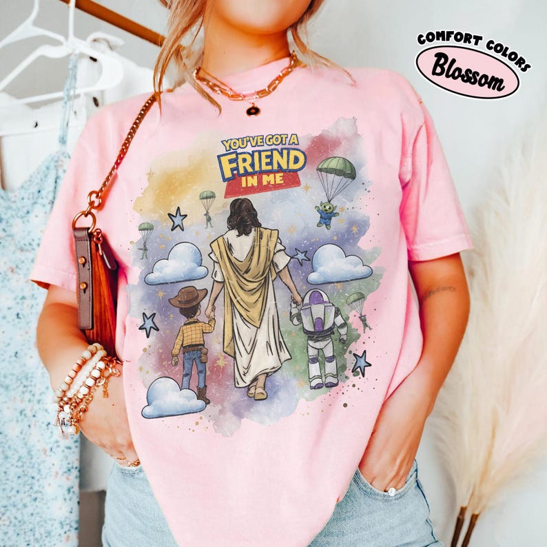 May include: Pink Comfort Colors t-shirt with a graphic featuring Jesus holding hands with cartoon characters. The text "YOU'VE GOT A FRIEND IN ME" is above the image. The shirt has a round neckline and short sleeves.