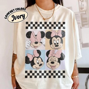 May include: Ivory-colored t-shirt featuring a four-panel design of Minnie Mouse faces with pink bows. The design is framed by a black and white checkered pattern. The words "COMFORT COLORS" and "Ivory" are printed on the upper left.