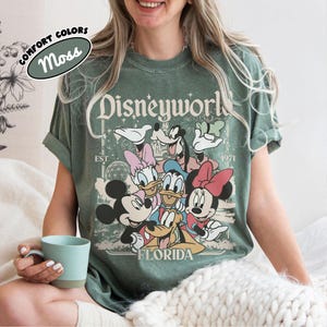 May include: Moss green t-shirt featuring a Disneyworld graphic with Mickey Mouse, Minnie Mouse, Donald Duck, Daisy Duck, Goofy, and Pluto. The shirt has the text "Disneyworld Florida EST. 1971" in a vintage style.