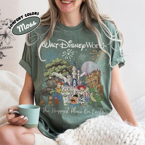 May include: Moss green t-shirt featuring a colorful graphic of Disney characters and landmarks, including a castle, train, and fireworks. The text "Walt Disney World" and "The Happiest Place On Earth" are printed on the shirt.