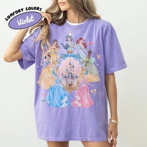 May include: A violet-colored t-shirt featuring a graphic of several cartoon princesses surrounding a castle. The shirt has short sleeves and a relaxed fit. The text "COMFORT COLORS" and "Violet" are printed on a purple oval.