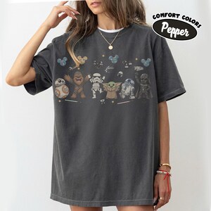 K&ouml;nnte beinhalten: Ein anthrazitfarbenes T-Shirt mit einem Grafikdruck von Star Wars-Figuren, darunter BB-8, Chewbacca, ein Stormtrooper, Grogu, R2-D2 und Darth Vader. Das Shirt zeigt kleine Sterne, Mickey-f&ouml;rmige Symbole und den Text Comfort Colors Pepper.