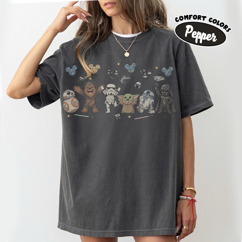 K&ouml;nnte beinhalten: Ein anthrazitfarbenes T-Shirt mit einem Grafikdruck von Star Wars-Figuren, darunter BB-8, Chewbacca, ein Stormtrooper, Grogu, R2-D2 und Darth Vader. Das Shirt zeigt kleine Sterne, Mickey-f&ouml;rmige Symbole und den Text Comfort Colors Pepper.