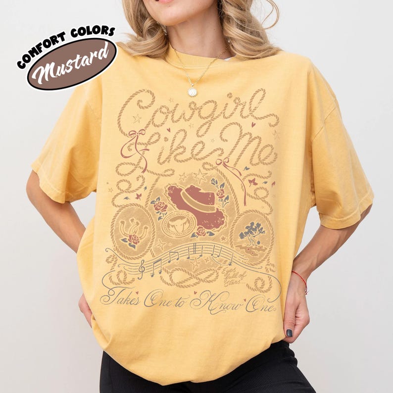 May include: A mustard yellow short-sleeve t-shirt featuring a brown graphic print with the text "Cowgirl Like Me" and "Takes One to Know One". The design includes a cowboy hat, a horseshoe, a tree, musical notes, and rope-style borders.