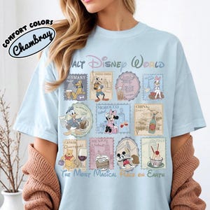 May include: Light blue t-shirt with a Walt Disney World graphic. The design features stamps with Disney characters and country names. The text "COMFORT COLORS Chambray" is on the upper left. The phrase "THE MOST MAGICAL PLACE ON EARTH" is at the bottom.