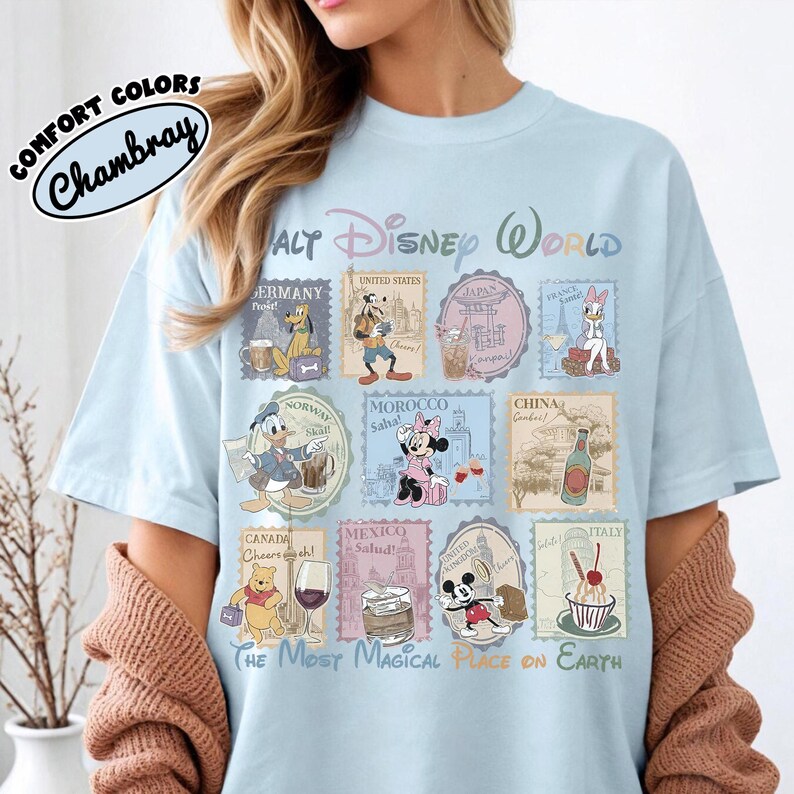 May include: Light blue t-shirt with a Walt Disney World graphic. The design features stamps with Disney characters and country names. The text "COMFORT COLORS Chambray" is on the upper left. The phrase "THE MOST MAGICAL PLACE ON EARTH" is at the bottom.