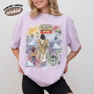 May include: A light purple t-shirt with the text "YOU'VE GOT A FRIEND IN ME" and a colorful graphic featuring cartoon characters and Jesus. The shirt also has the words "COMFORT COLORS" and "orchid" printed on it.