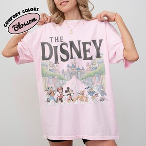 Może przedstawiać: Jasnor&oacute;żowa koszulka z czarnym napisem "THE DISNEY". Koszulka przedstawia grafikę postaci Disneya przechodzących przez ulicę z zamkiem w tle. Na koszulce znajdują się r&oacute;wnież słowa "COMFORT COLORS" i "Blossom".