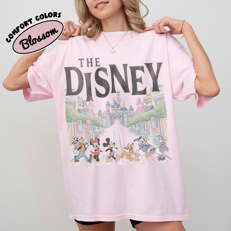 Może przedstawiać: Jasnor&oacute;żowa koszulka z czarnym napisem "THE DISNEY". Koszulka przedstawia grafikę postaci Disneya przechodzących przez ulicę z zamkiem w tle. Na koszulce znajdują się r&oacute;wnież słowa "COMFORT COLORS" i "Blossom".
