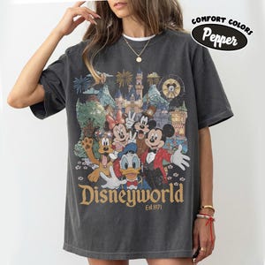 May include: Dark gray t-shirt featuring a colorful graphic of Disney characters, including Mickey Mouse, Minnie Mouse, and Donald Duck, in front of a castle and fireworks display. The text "Disneyworld Est. 1971" is printed below the graphic. The shirt has a Comfort Colors Pepper label.