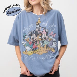 May include: Blue t-shirt with a graphic of Disney characters including Mickey Mouse, Donald Duck, Goofy, Pluto, Minnie Mouse, and Daisy Duck. The shirt has the words "Comfort Colors" and "Blue Jean" in a circular design.