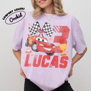Puede incluir: Una camiseta de color lila claro con un gr&aacute;fico de un coche de carreras rojo, banderas a cuadros y el n&uacute;mero 3. El nombre "LUCAS" est&aacute; impreso en rojo debajo del coche. La camiseta tambi&eacute;n tiene el texto "COMFORT COLORS" y "Orchid".