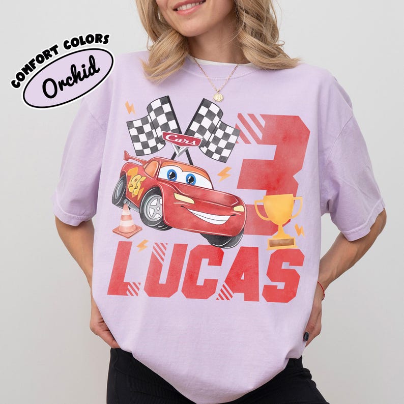 Puede incluir: Una camiseta de color lila claro con un gr&aacute;fico de un coche de carreras rojo, banderas a cuadros y el n&uacute;mero 3. El nombre "LUCAS" est&aacute; impreso en rojo debajo del coche. La camiseta tambi&eacute;n tiene el texto "COMFORT COLORS" y "Orchid".