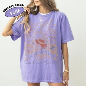 May include: A violet short-sleeve t-shirt featuring a graphic print with the text Cowgirl Like Me and Takes One to Know One. The design includes a cowboy hat, musical notes, and floral elements in orange, red, and blue tones on the front.