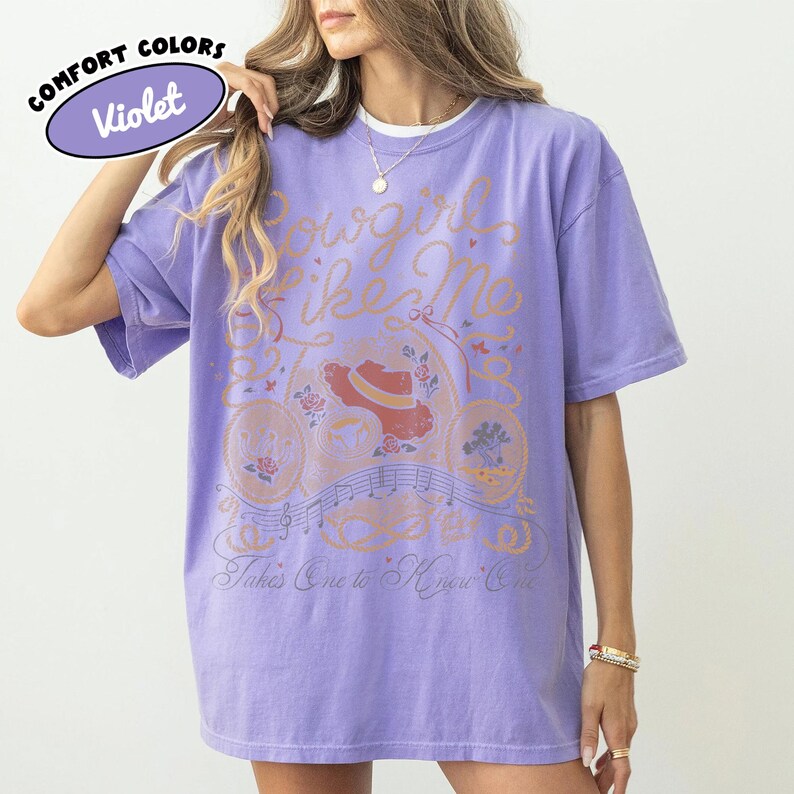 May include: A violet short-sleeve t-shirt featuring a graphic print with the text Cowgirl Like Me and Takes One to Know One. The design includes a cowboy hat, musical notes, and floral elements in orange, red, and blue tones on the front.