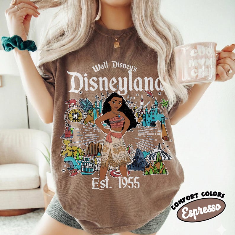 Op de afbeelding: Een bruin T-shirt met korte mouwen en een grafische print van Moana in het midden, omringd door diverse Disneyland-parkiconen. De tekst luidt Walt Disney's Disneyland Est. 1955. Het shirt heeft een ronde hals en een relaxte pasvorm.