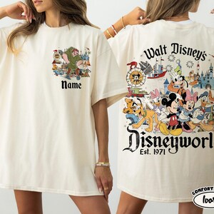 May include: Ivory-colored t-shirt featuring a Disney-themed graphic. The front displays a cartoon image with the text "Name". The back showcases Disney characters with "Walt Disney's Disneyworld Est. 1971".