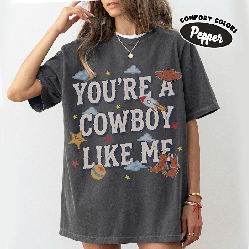 May include: A charcoal gray short-sleeve t-shirt featuring the text YOU'RE A COWBOY LIKE ME in white lettering. The design includes illustrations of a brown cowboy hat, a rocket, a sheriff star, a ball, and cowboy boots, along with blue clouds and stars.