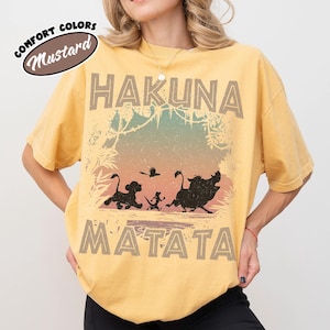 May include: Mustard-coloured t-shirt with the words "HAKUNA MATATA" in a distressed font. The shirt features silhouettes of Simba, Timon, Pumbaa, and a bird. The words "Comfort Colors" and "Mustard" are in an oval.