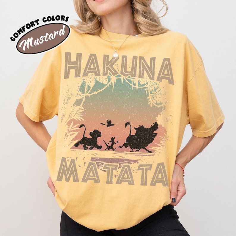 May include: Mustard-coloured t-shirt with the words "HAKUNA MATATA" in a distressed font. The shirt features silhouettes of Simba, Timon, Pumbaa, and a bird. The words "Comfort Colors" and "Mustard" are in an oval.