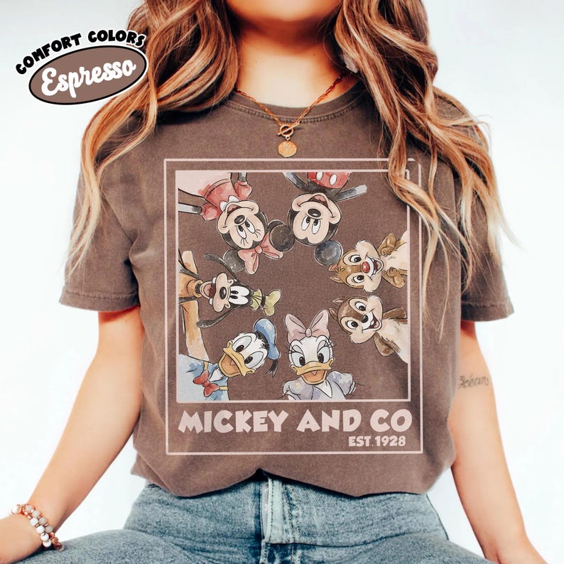 Puede incluir: Camiseta color espresso con un gr&aacute;fico enmarcado de Mickey Mouse, Minnie Mouse, Donald Duck, Daisy Duck, Goofy y Chip y Chop. El texto "Mickey and Co. EST. 1928" est&aacute; impreso debajo. La camiseta tiene mangas cortas.