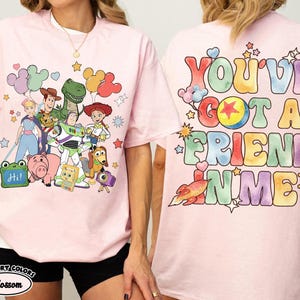 May include: A light pink t-shirt featuring a front graphic of various animated characters including a cowboy, space ranger, cowgirl, dinosaur, and pig, with balloons and a sign saying Hi. The back displays the text You've Got A Friend In Me.