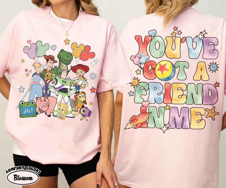 May include: A light pink t-shirt featuring a front graphic of various animated characters including a cowboy, space ranger, cowgirl, dinosaur, and pig, with balloons and a sign saying Hi. The back displays the text You've Got A Friend In Me.