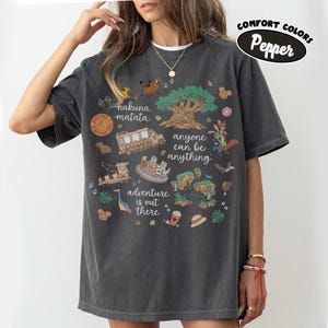 Puede incluir: Camiseta Comfort Colors gris oscuro con un dise&ntilde;o gr&aacute;fico colorido. El dise&ntilde;o incluye los textos "nakuna matata", "anyone can be anything" y "adventure is out there". La camiseta presenta varias ilustraciones de dibujos animados.