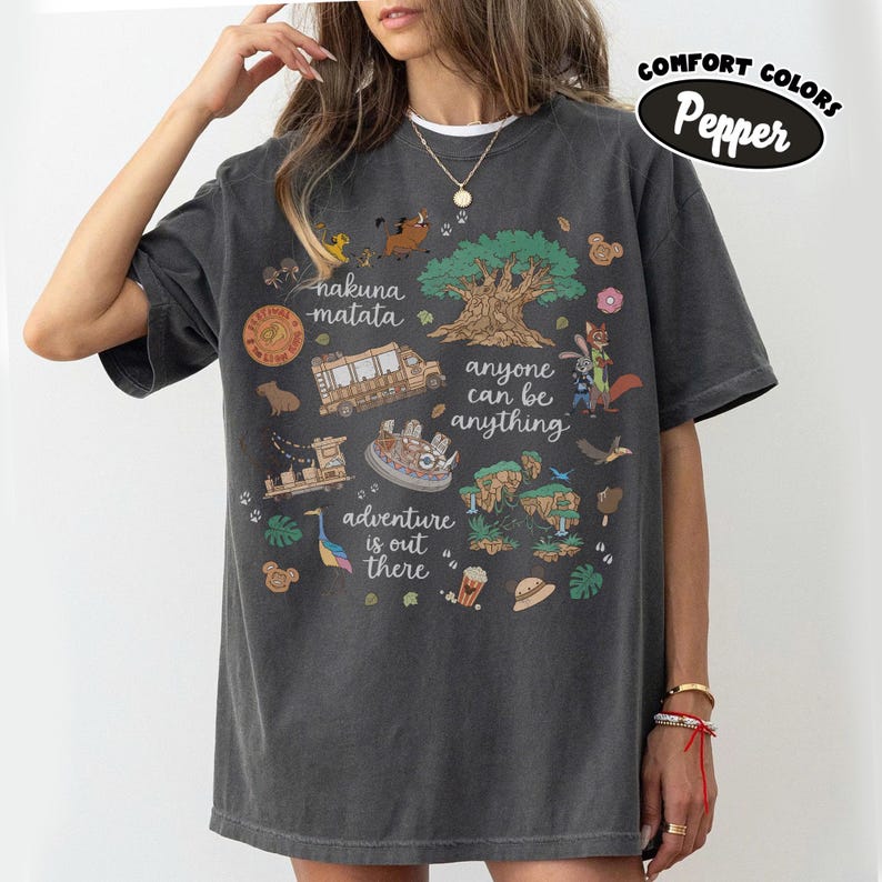 Puede incluir: Camiseta Comfort Colors gris oscuro con un dise&ntilde;o gr&aacute;fico colorido. El dise&ntilde;o incluye los textos "nakuna matata", "anyone can be anything" y "adventure is out there". La camiseta presenta varias ilustraciones de dibujos animados.