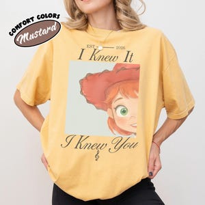 Op de afbeelding: Een mosterdgeel T-shirt met korte mouwen en een afbeelding van een stripfiguur met rood haar en een rode hoed. De tekst luidt "EST 2026 I Knew It I Knew You" met een muzieknoot. Het shirt heeft een ronde hals en een relaxte pasvorm.