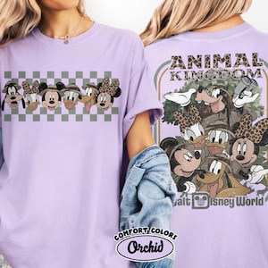 K&ouml;nnte beinhalten: Ein helllila T-Shirt mit einer Grafik von Disney-Charakteren, darunter Mickey, Minnie, Donald, Daisy, Goofy und Pluto, mit Safarih&uuml;ten. Auf der R&uuml;ckseite des Shirts steht der Text "ANIMAL KINGDOM" und weitere Charaktere. Das Shirt ist mit "COMFORT COLORS Orchid" gekennzeichnet.
