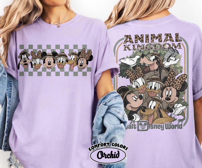 K&ouml;nnte beinhalten: Ein helllila T-Shirt mit einer Grafik von Disney-Charakteren, darunter Mickey, Minnie, Donald, Daisy, Goofy und Pluto, mit Safarih&uuml;ten. Auf der R&uuml;ckseite des Shirts steht der Text "ANIMAL KINGDOM" und weitere Charaktere. Das Shirt ist mit "COMFORT COLORS Orchid" gekennzeichnet.