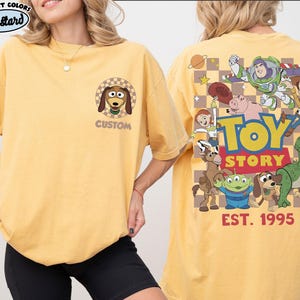 Puede incluir: Camiseta amarilla mostaza con una imagen de dibujos animados de Slinky Dog en la parte delantera y personajes de Toy Story en la parte trasera. La parte trasera de la camiseta tiene las palabras "TOY STORY" y "EST. 1995". La camiseta es de la marca Comfort Colors.