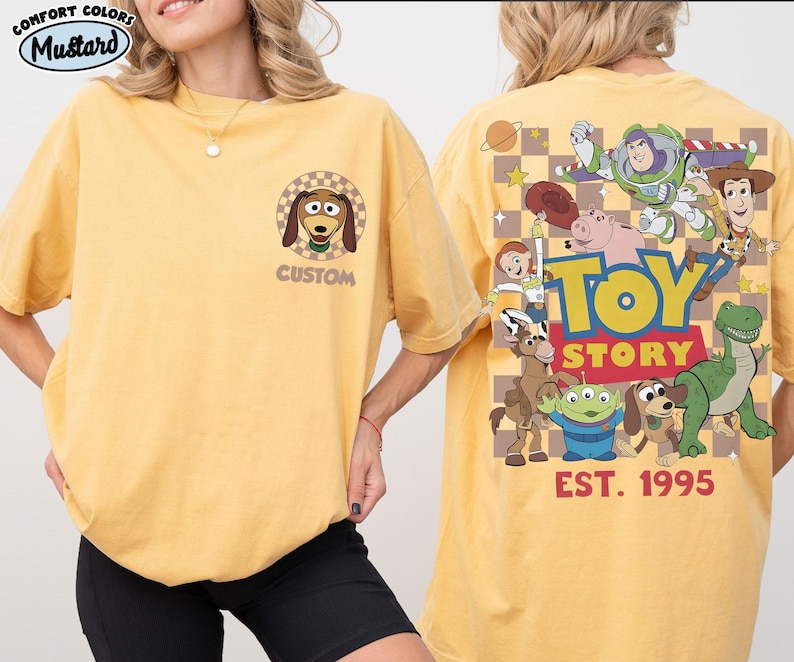 Puede incluir: Camiseta amarilla mostaza con una imagen de dibujos animados de Slinky Dog en la parte delantera y personajes de Toy Story en la parte trasera. La parte trasera de la camiseta tiene las palabras "TOY STORY" y "EST. 1995". La camiseta es de la marca Comfort Colors.