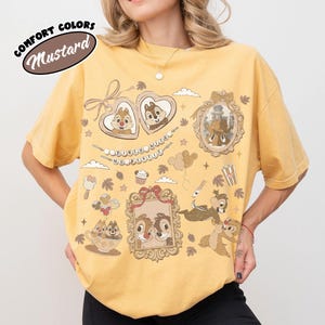 May include: Mustard-colored t-shirt with a graphic design featuring cartoon characters in frames and decorative elements. The shirt has short sleeves and a crew neck. The text "Comfort Colors" and "Mustard" are printed on the upper left side of the shirt.