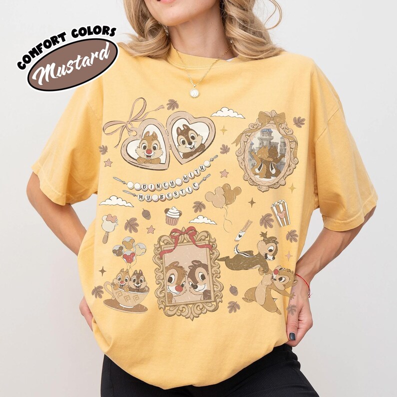 May include: Mustard-colored t-shirt with a graphic design featuring cartoon characters in frames and decorative elements. The shirt has short sleeves and a crew neck. The text "Comfort Colors" and "Mustard" are printed on the upper left side of the shirt.