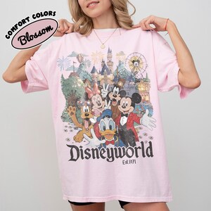 May include: A light pink t-shirt featuring a colorful graphic of Disney characters, including Mickey Mouse, Minnie Mouse, and Donald Duck. The word "Disneyworld" is printed in a vintage font, with "Est.1971" below. The shirt also has the words "Comfort Colors" and "Blossom" printed on it.