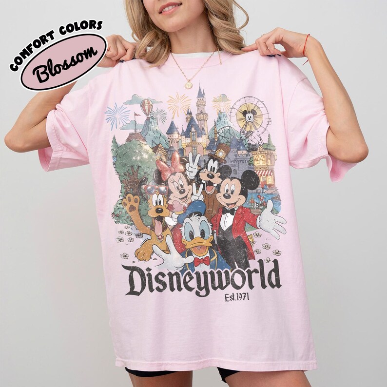 May include: A light pink t-shirt featuring a colorful graphic of Disney characters, including Mickey Mouse, Minnie Mouse, and Donald Duck. The word "Disneyworld" is printed in a vintage font, with "Est.1971" below. The shirt also has the words "Comfort Colors" and "Blossom" printed on it.
