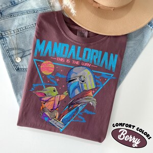 Może przedstawiać: Koszulka w kolorze jagodowym z grafiką The Mandalorian i Grogu, z napisem "MANDALORIAN - THIS IS THE WAY" w kolorze niebieskim. Koszulka jest częścią kolekcji Comfort Colors. Beżowy kapelusz i niebieskie dżinsy w tle.