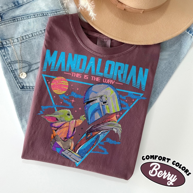 Może przedstawiać: Koszulka w kolorze jagodowym z grafiką The Mandalorian i Grogu, z napisem "MANDALORIAN - THIS IS THE WAY" w kolorze niebieskim. Koszulka jest częścią kolekcji Comfort Colors. Beżowy kapelusz i niebieskie dżinsy w tle.