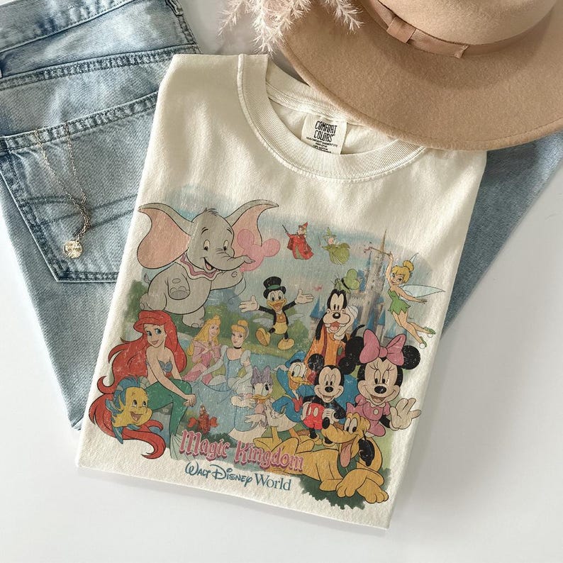 May include: Cream-colored t-shirt featuring a colorful graphic of Disney characters, including Dumbo, Ariel, Mickey Mouse, and Minnie Mouse. The text "Magic Kingdom Walt Disney World" is printed below the characters. The shirt is folded on top of blue jeans.