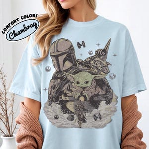 Puede incluir: Camiseta azul claro con un dise&ntilde;o de The Mandalorian, Grogu y una nave espacial. El dise&ntilde;o es en tonos grises y marrones, con estrellas y elementos espaciales. El texto "COMFORT COLORS Chambray" est&aacute; impreso en la parte superior izquierda.