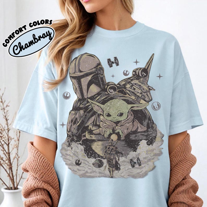 Puede incluir: Camiseta azul claro con un dise&ntilde;o de The Mandalorian, Grogu y una nave espacial. El dise&ntilde;o es en tonos grises y marrones, con estrellas y elementos espaciales. El texto "COMFORT COLORS Chambray" est&aacute; impreso en la parte superior izquierda.