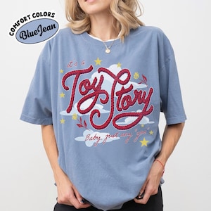 May include: A blue jean colored short-sleeve t-shirt featuring a graphic print with the red rope-style text "it's a Toy Story" and "Baby, just say yes". The design includes white clouds, yellow stars, and small red flowers on a blue background.