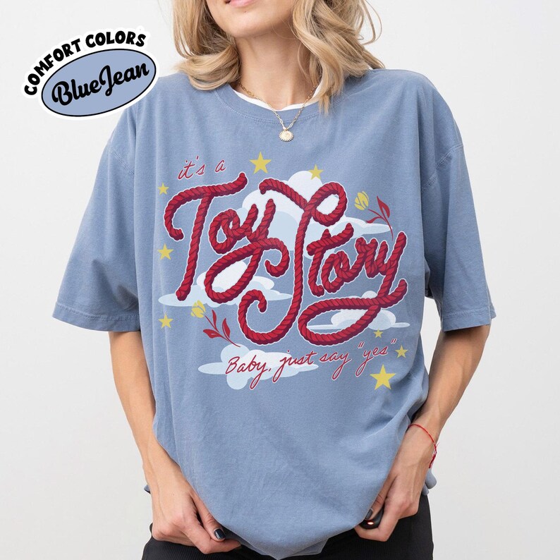 May include: A blue jean colored short-sleeve t-shirt featuring a graphic print with the red rope-style text "it's a Toy Story" and "Baby, just say yes". The design includes white clouds, yellow stars, and small red flowers on a blue background.