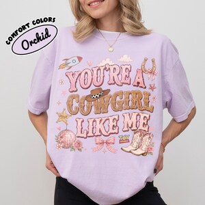 Puede incluir: Una camiseta de color morado claro con el texto "YOU'RE A COWGIRL LIKE ME" en rosa y marr&oacute;n. El gr&aacute;fico incluye un cohete, un sombrero de vaquero, una herradura, una estrella de sheriff, una bola de discoteca, un lazo y botas con el logo "TOY STORY".