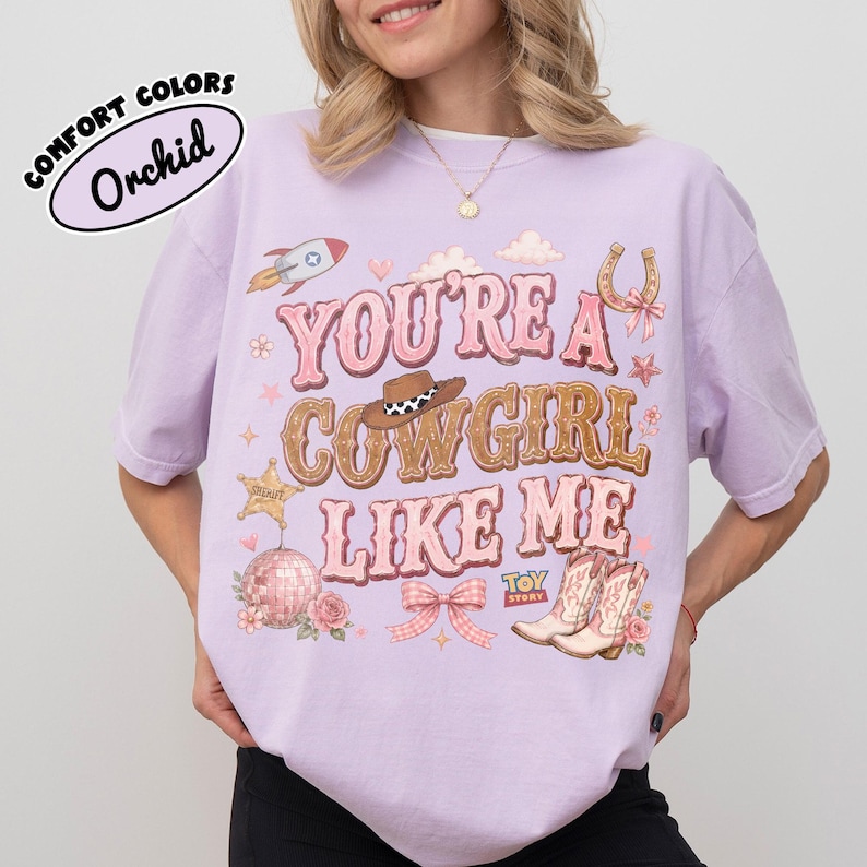 Puede incluir: Una camiseta de color morado claro con el texto "YOU'RE A COWGIRL LIKE ME" en rosa y marr&oacute;n. El gr&aacute;fico incluye un cohete, un sombrero de vaquero, una herradura, una estrella de sheriff, una bola de discoteca, un lazo y botas con el logo "TOY STORY".